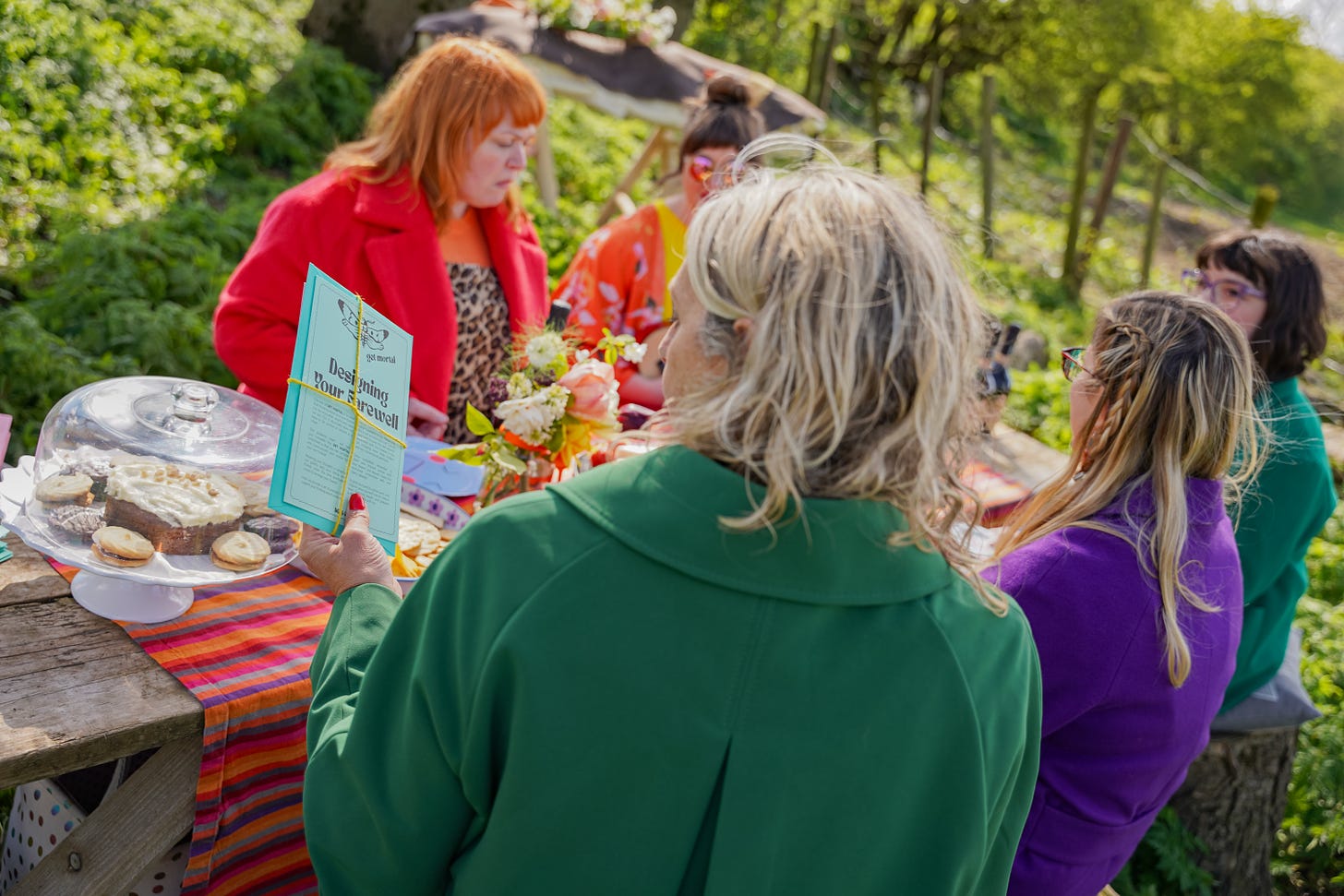 five brightly dressed white women sitting at a picnic table in a woodland burial ground with a shroud in the background