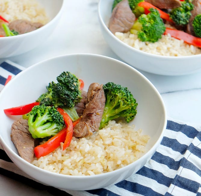 Beef & Broccoli stir fry, with sliced red peppers, on top of brown rice, sits in a white bowl, on a striped blue and white towel on top of a white table. 