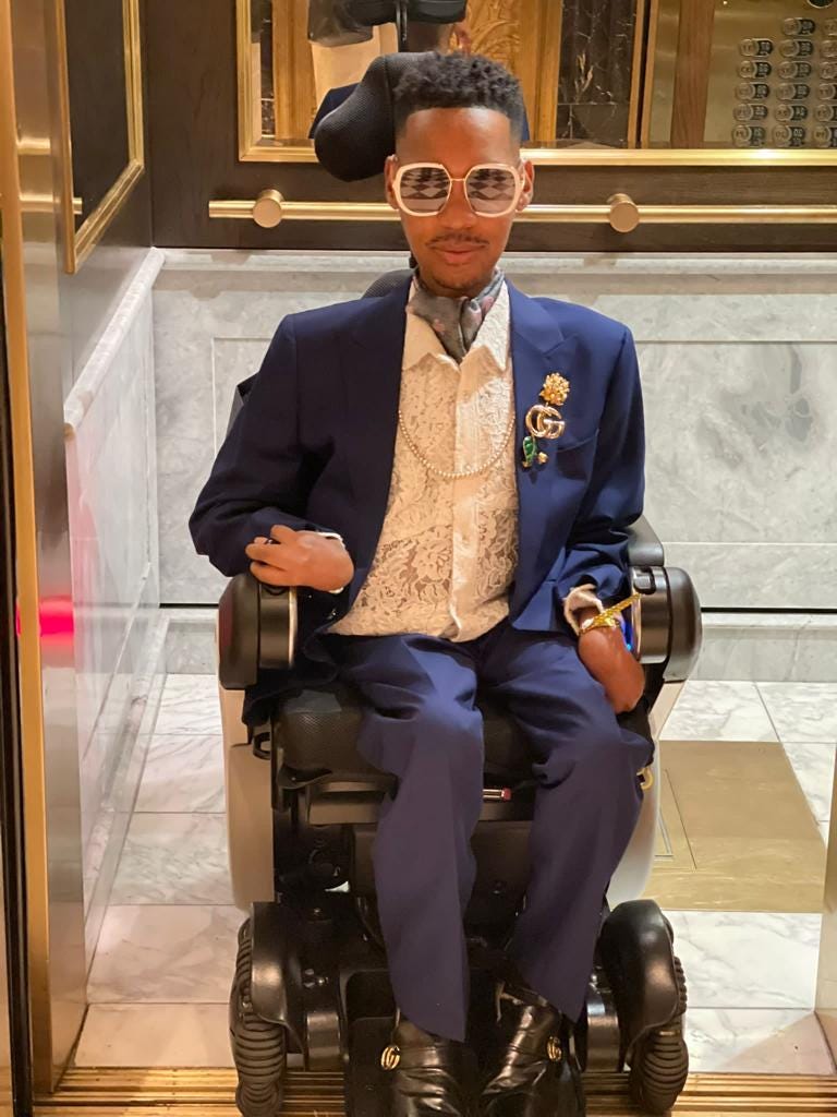 Eddie Ndopu, a black man in a power wheelchair, rolling out of an elevator wearing a dark blue Gucci suit, white lace shirt, gray ascot and large white-framed sunglasses.