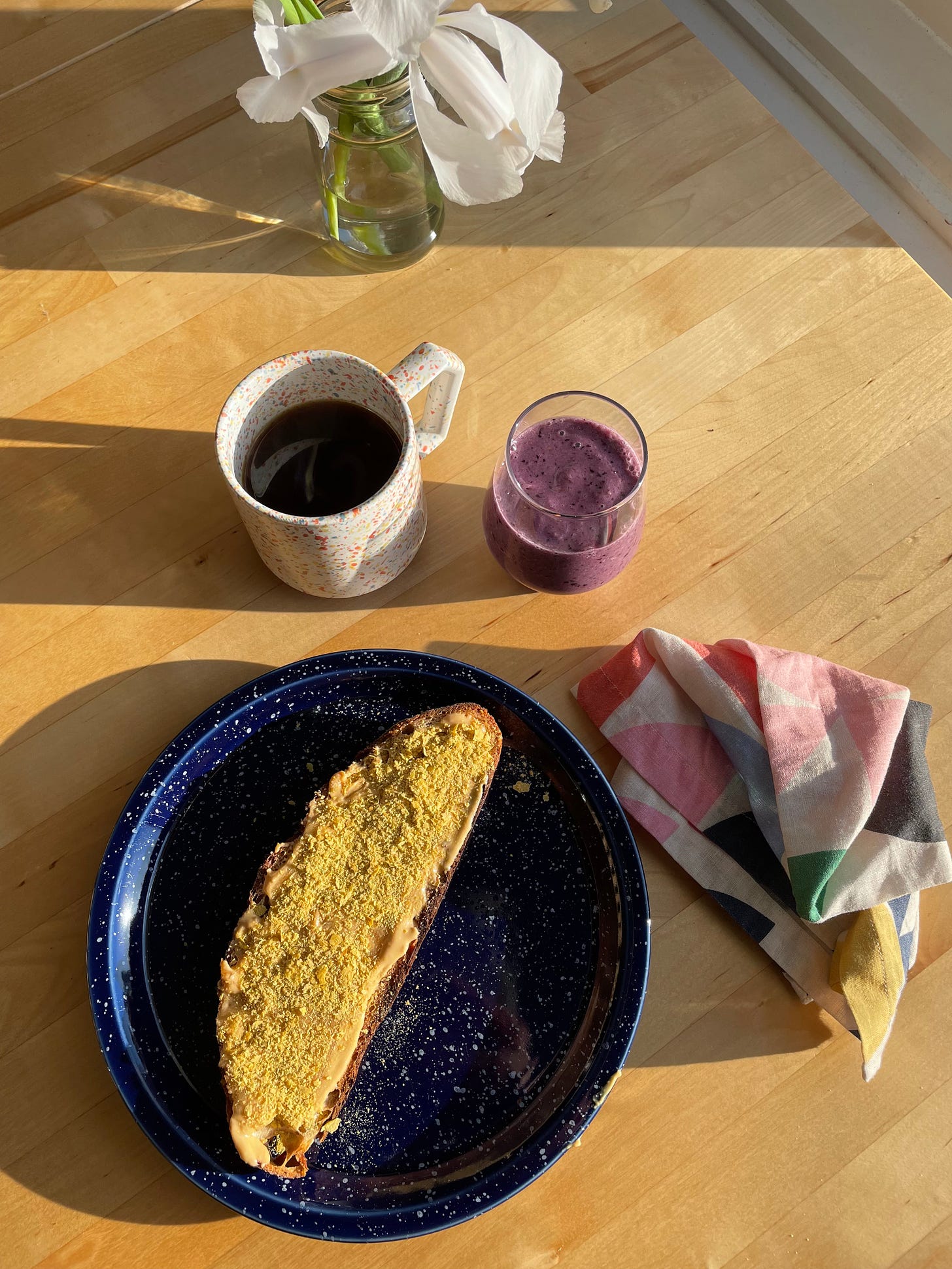 Piece of toast on a speckled plate alongside a cup of black coffee and a glass of blueberry smoothie