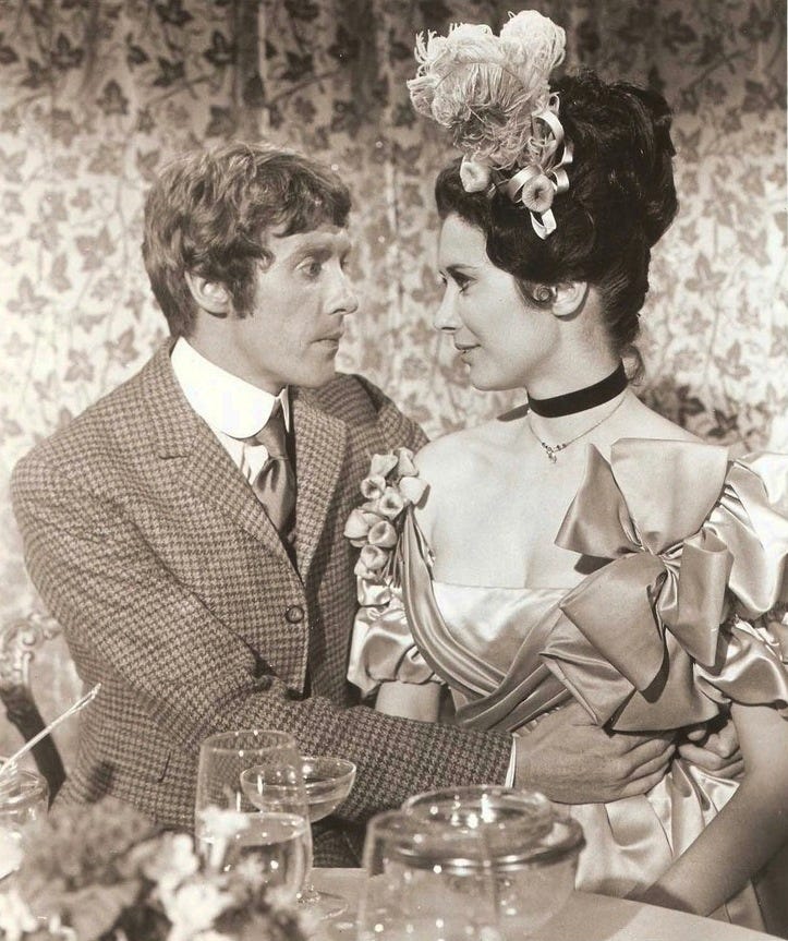 a nervous blonde white man in a tweed suit and pretty brunette white woman in a turn of the century style dress and hairdo look at each other pointedly.
