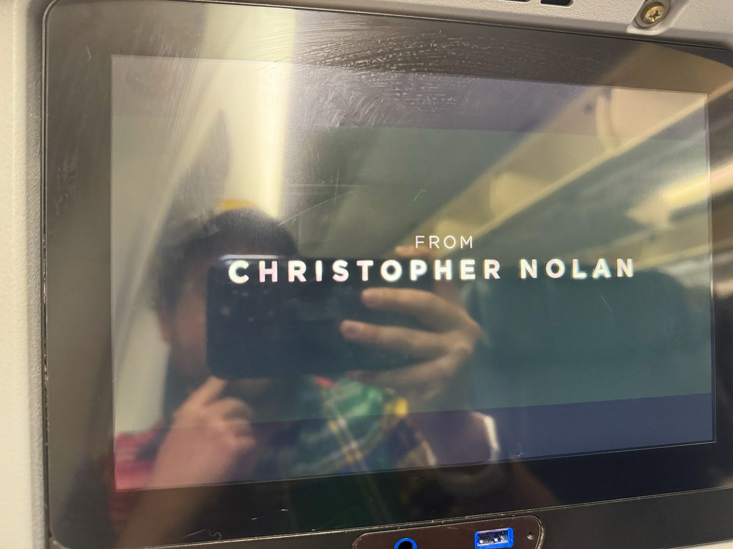 Photograph of an aircraft seatback screen with the words "FROM CHRISTOPHER NOLAN" and the reflection of a man taking a picture using a smartphone.