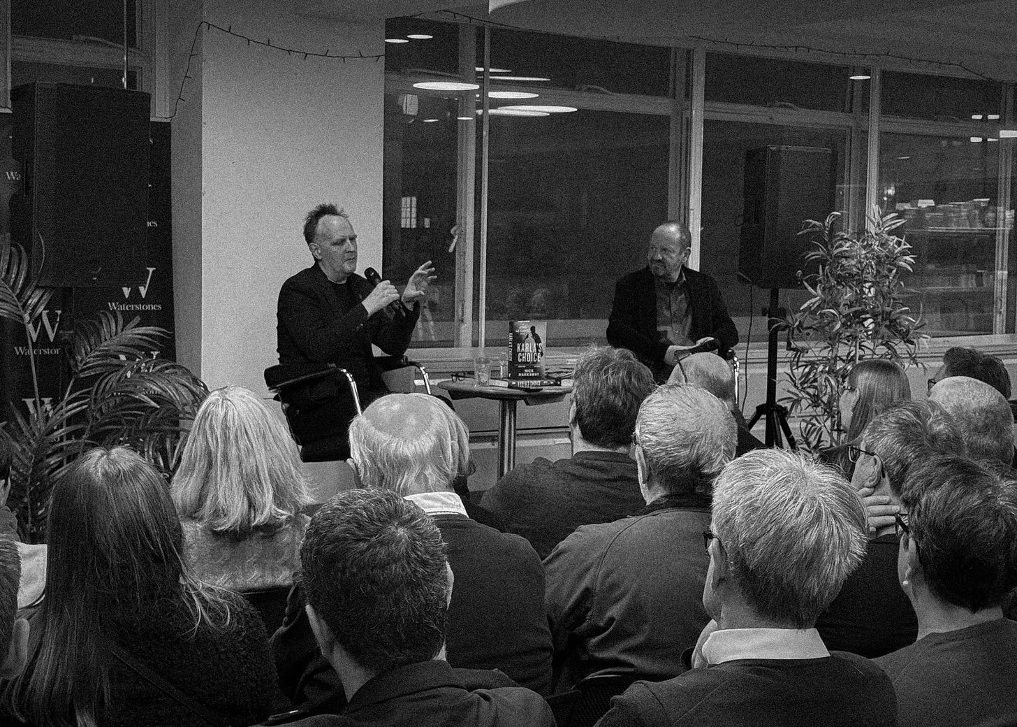 Black and white photograph shows a man wearing black speaking into a microphone. Compere and audience looks on.