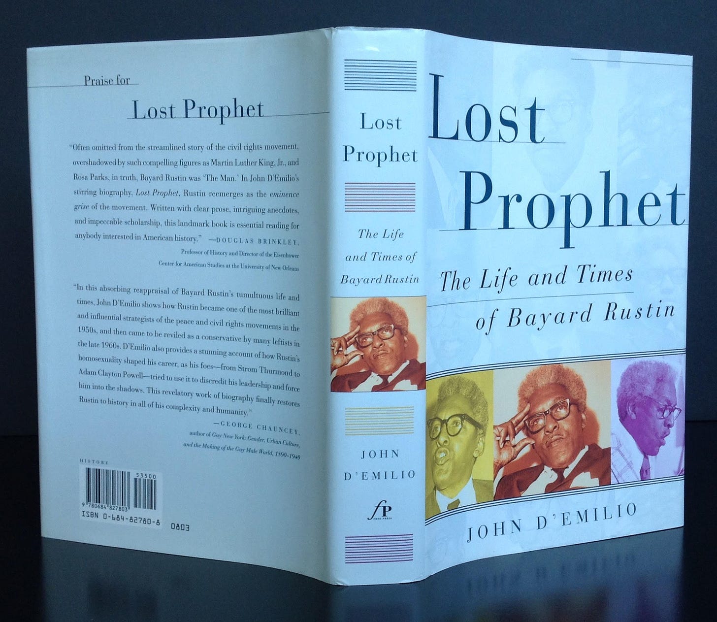 The photo shows a book split open, propped up, and with the book cover facing the camera. The left side shows the back cover of the book showing text "Praise for Lost Prophet" followed by small text in two paragraphs. The book spine's text reads, "Lost Prophet: The Life and Times of Bayard Rustin, Joh D'Emilio". The right side of the photo shows the front cover of the book with text that reads, "Lost Prophet: The Life and Times of Bayard Rustin, John D'Emilio" with yellow, tan, and purple photos of Bayard Rustin in a row, side-by-side.