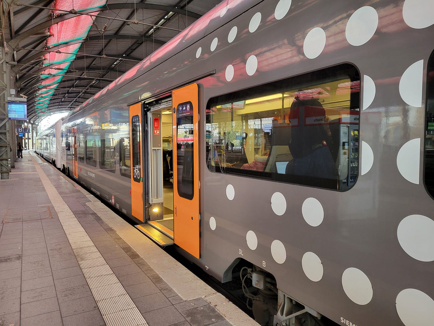 Gray metal passenger train with white dots and orange doors sits at a platform in the station.