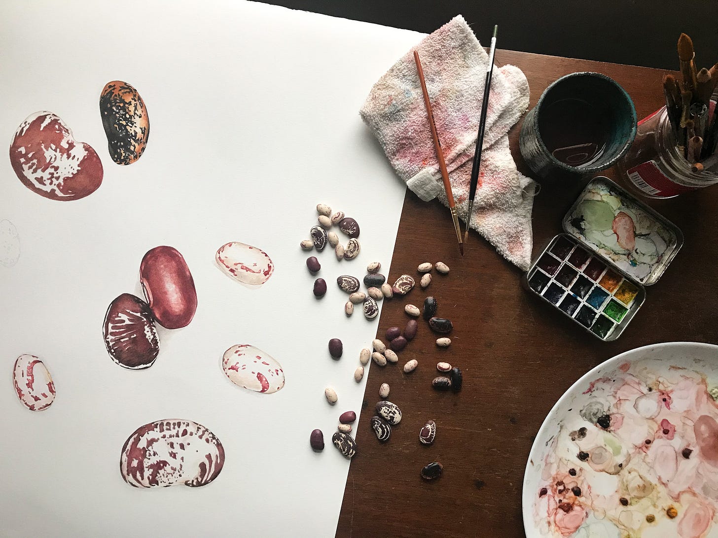 A snap of the painting as it is being painted. Dried beans lay on top of the larger painting, with palette and brushes to the right 