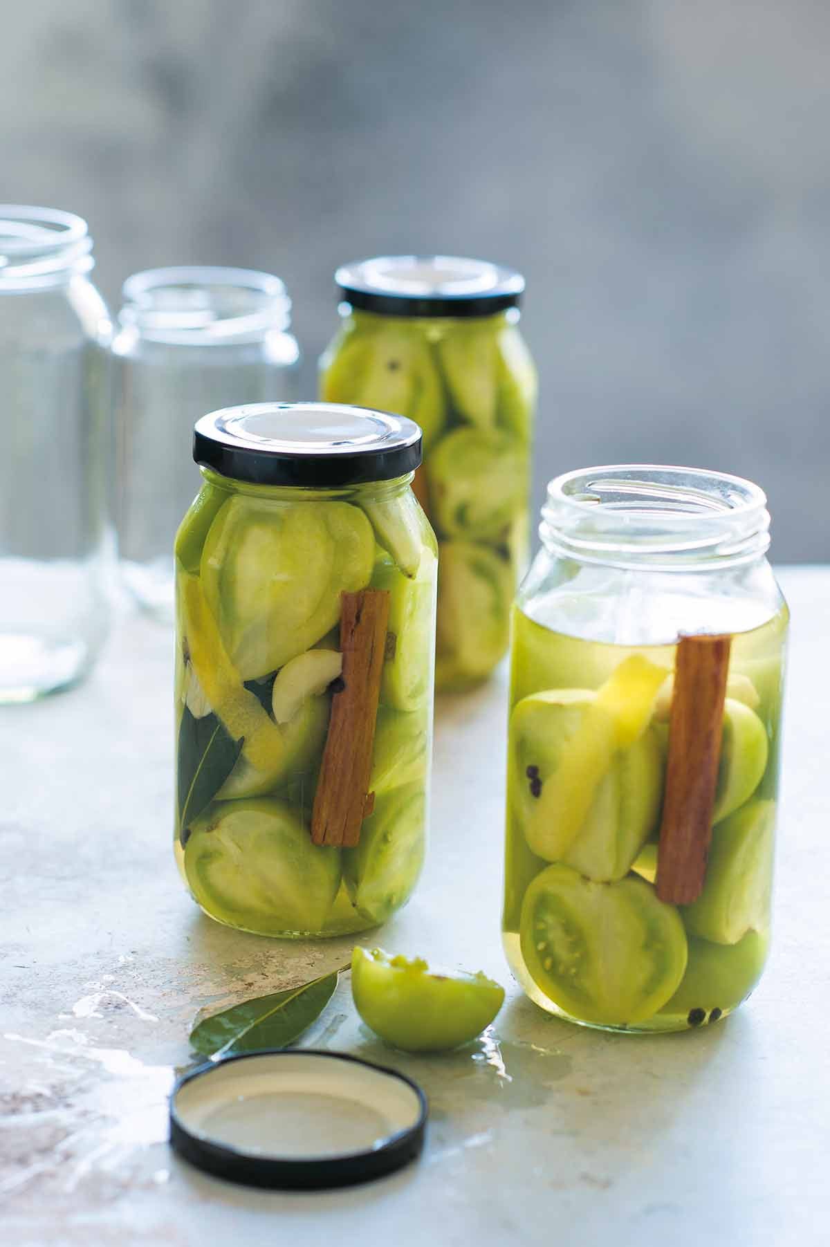 Three jars filled with pickled green tomatoes and a cinnamon stick with two empty jars in the background.