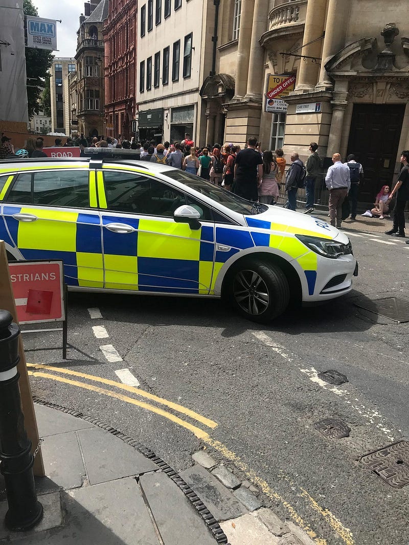 A police car, with a yellow-and-blue Battenberg pattern design, is closing off Clare Street in Bristol city centre to protect Dr Who filming. A crowd watches the filming taking place.