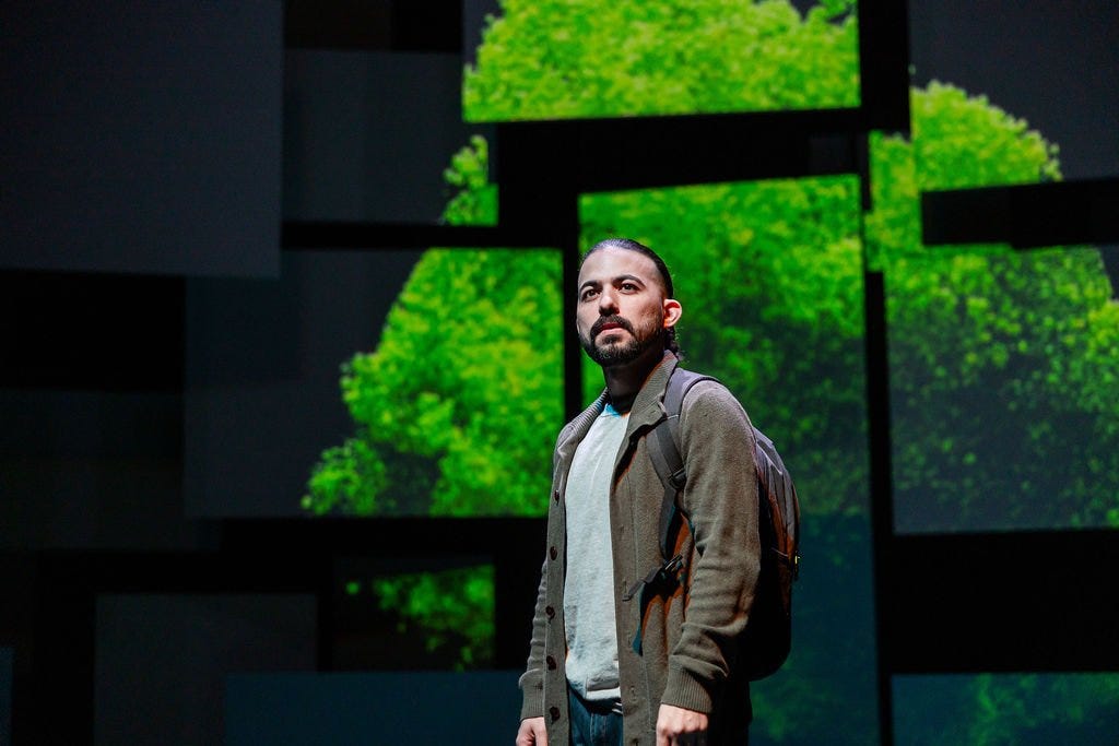 A man with dark hair in a ponytail and a dark beard, wearing a green jacket over a grey t-shirt, jeans, and a backpack, stares into the distance in front of multiple panels in which a green mass of spheres is projected
