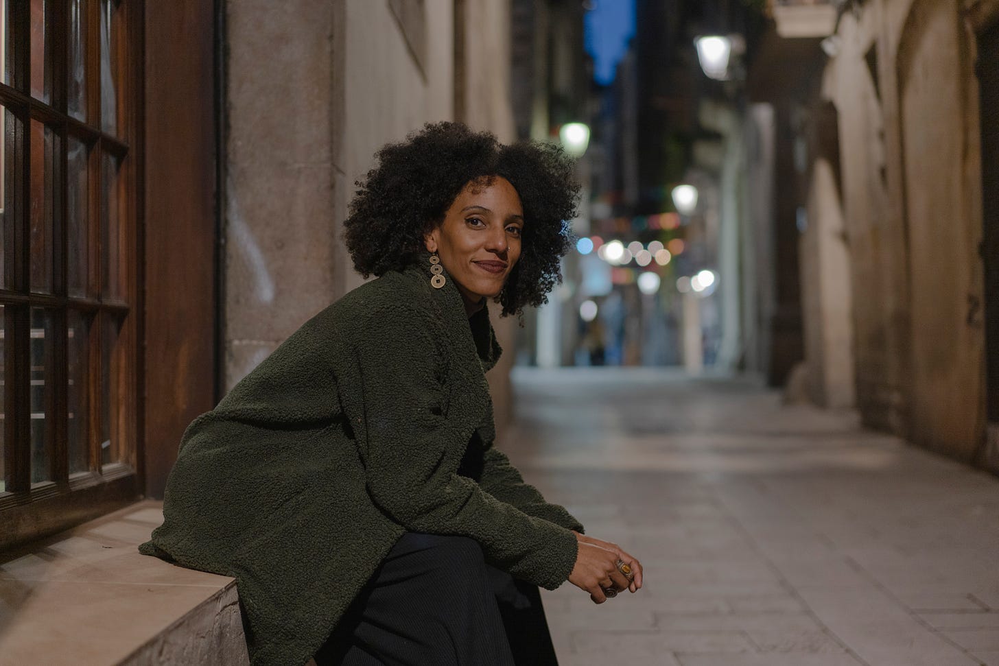 a black woman with dark curly hair sits on the ledge of a wooden window in an alley of Barcelona at dusk. She looks at the camera with a confident smile