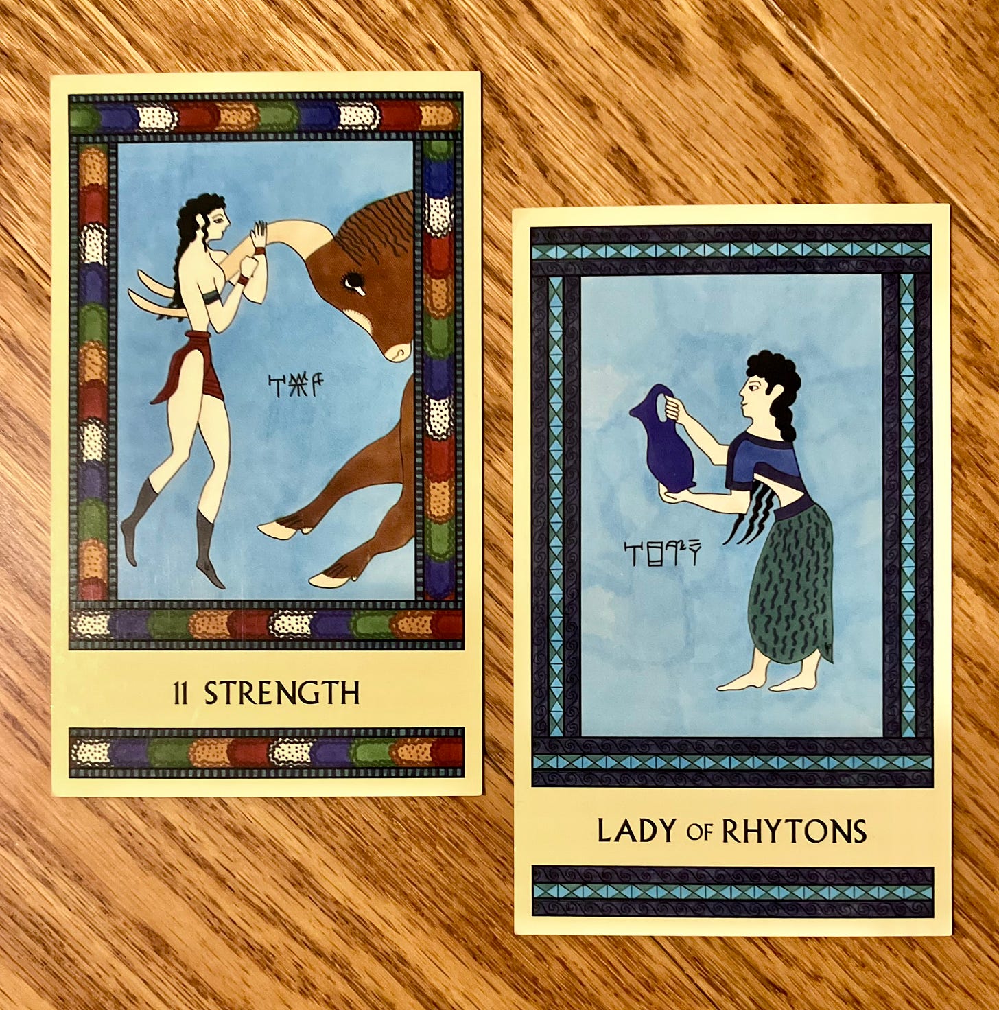 Two Minoan Tarot cards side by side on a deep golden wood surface. The Strength card has a multi-colored border. It shows a bull leaper hanging bodily from the horns of a galloping bull. The Lady of Rhytons is painted in shades of blue. It shows a Minoan woman standing, facing left, tipping a large pitcher as if she is about to pour from it.