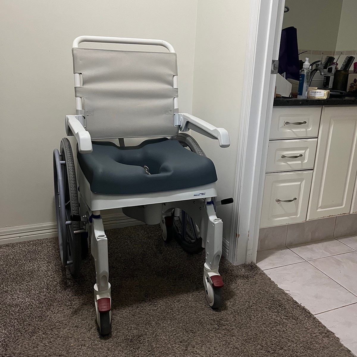 A grey and white combination of a toilet with bedpan and a manual wheelchair.