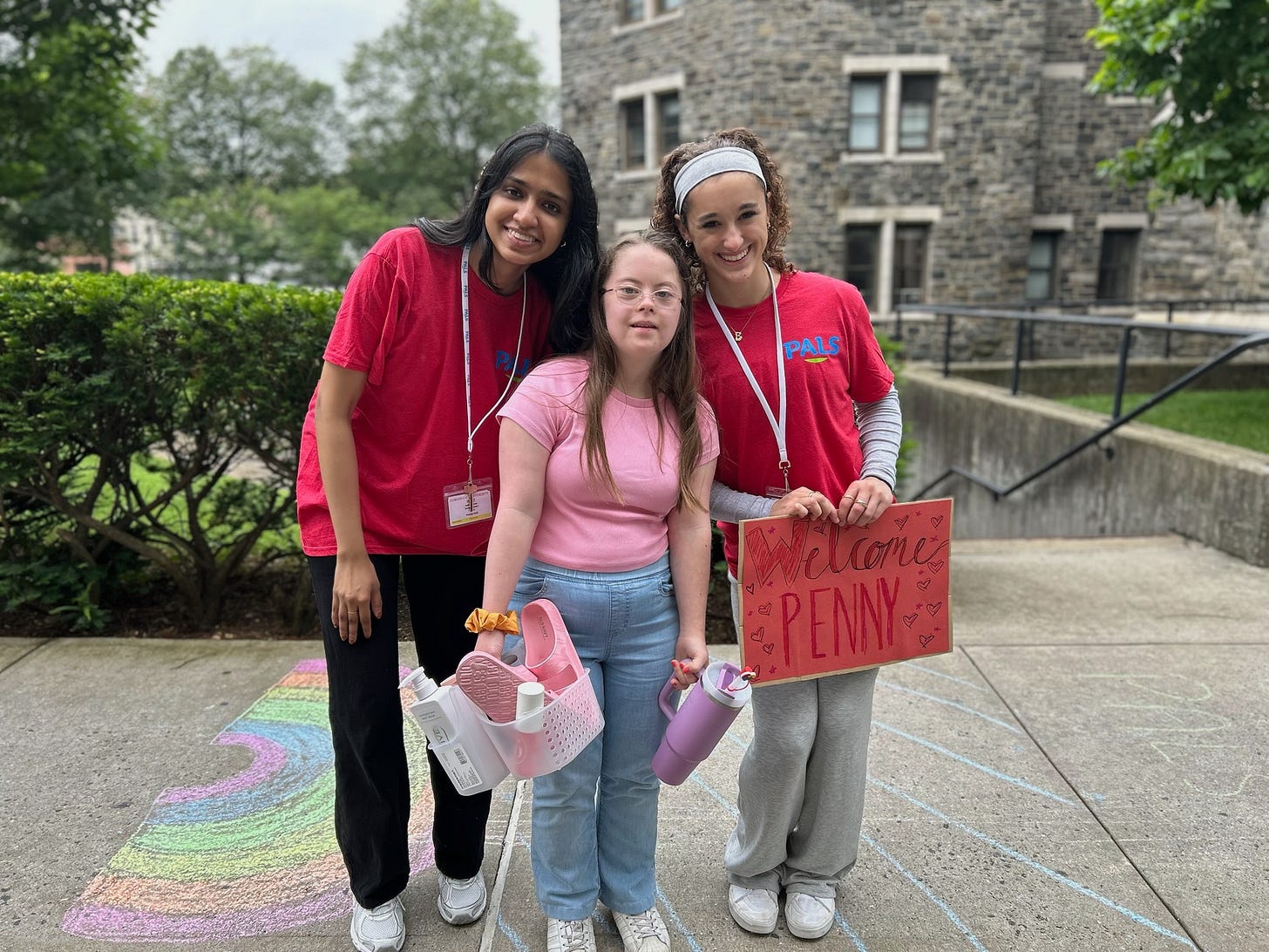 Penny and two members of a welcome team smile outdoors in front of a stone building. The welcome team is wearing matching red "PALS" shirts and lanyards, one of whom holds a handmade red sign that reads “Welcome Penny” with hearts. Penny and two members of a welcome team smile outdoors in front of a stone building. The welcome team is wearing matching red "PALS" shirts and lanyards, one of whom holds a handmade red sign that reads “Welcome Penny” with hearts.