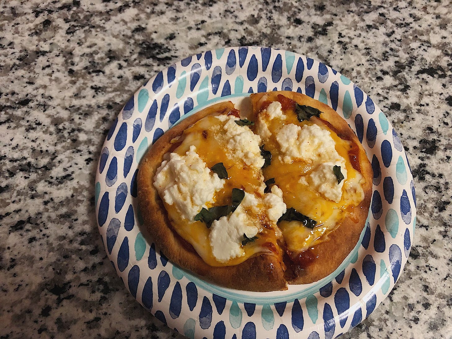 A overhead view of a mini naan pizza on a paper plate with blobs of ricotta and basil