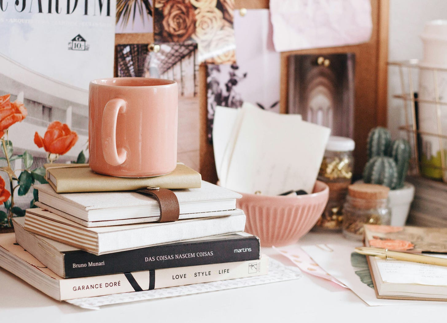 A desk is piled with books, a pink mug, pens and notebooks, and decorated with floral photos.