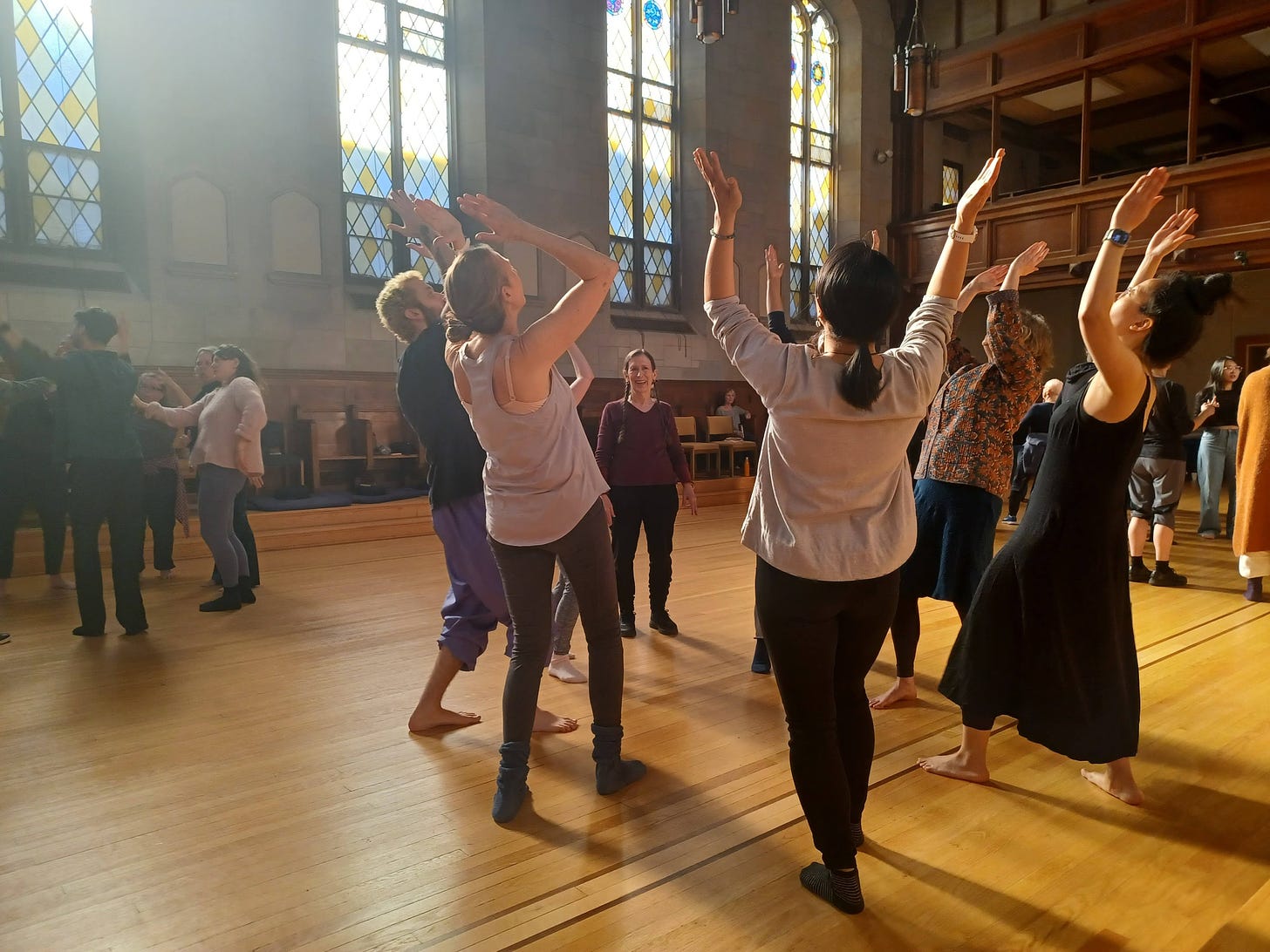 People are spread out in clusters in a large former chapel with stained glass windows and a golden wood floor. In the foreground Meredith Monk observes a circle of five people who are all leaning back with their arms in the air.