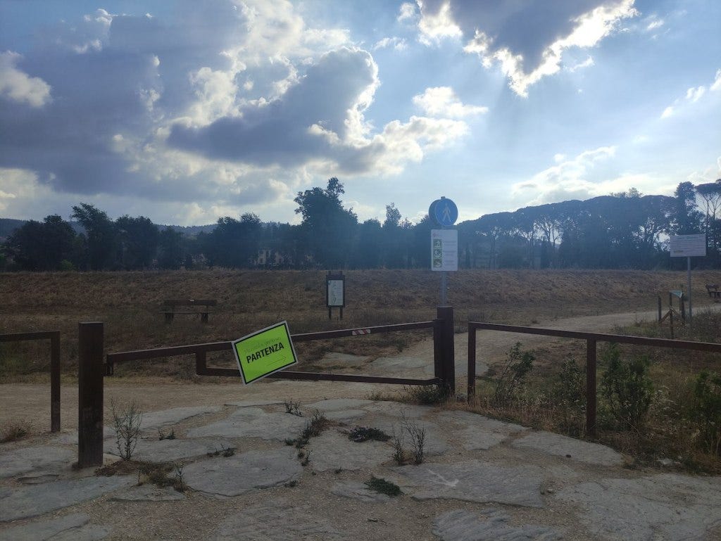 A sign at the park entrance, hanging on a metal gate, points participants to the start. Behind is a grassy bank, brown for lack of rain, with trees standing up behind that. A sign at the park entrance, hanging on a metal gate, points participants to the start. Behind is a grassy bank, brown for lack of rain, with trees standing up behind that.