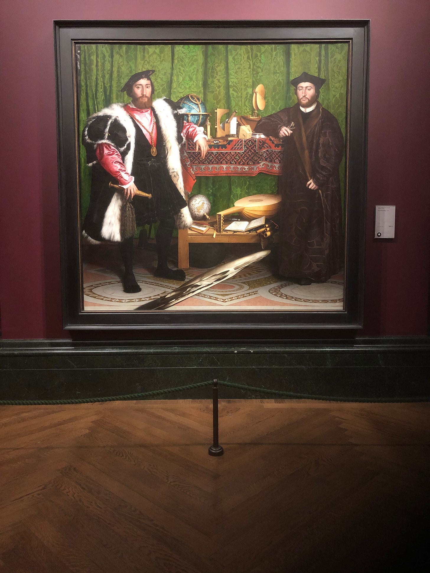 A painting of two C16th men, standing before a table and a green damask curtain. The men are well dressed and clearly of some importance. Various items are placed upon the table - a globe, mathematical instruments, a lute and sheet music. A strange image at the front of the painting is a distorted skull.
