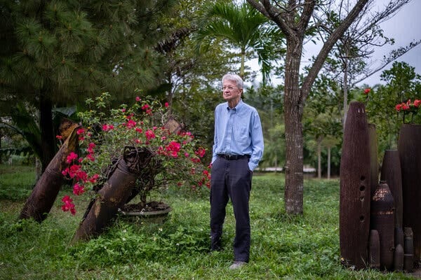 A tall, lanky man stands next to two deactivated bombs from which a profusion of red flowers pop out. 