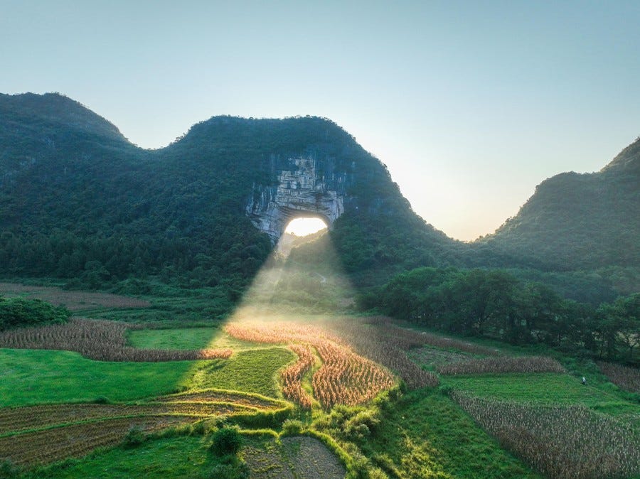 A sunbeam shines through a natural arch in a mountain, lighting up a field below.