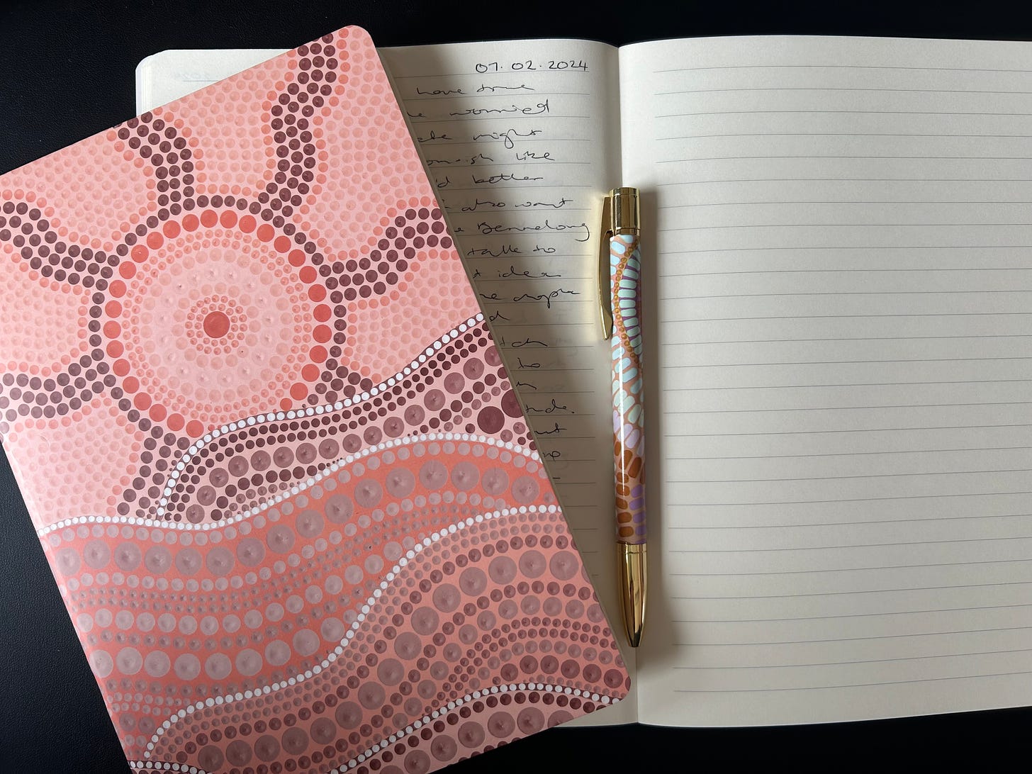 Two notebooks, one with the cover showing an Aboriginal dot painting design, the other open underneath partially showing some writing.