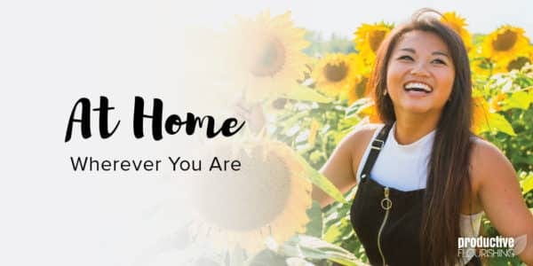 A woman with brown hair looks up and smiles while standing among sunflowers. Text Overlay: At Home Wherever You Are