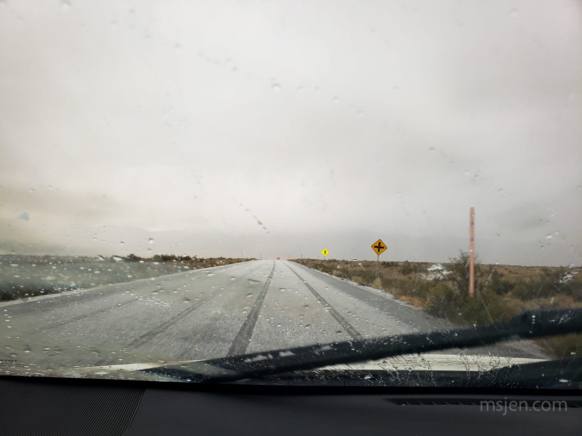 Driving down a country road with an inch of snow and a pair of tire tracks in front of the car with the camera. Snow on the sagebrush and foggy front car window. 