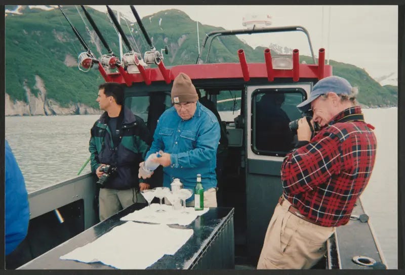 Three men stand in the back of a fishing boat with mountains in the background. On a table in the center, now-dead villain of American legal policy Antonin Scalia pours vodka into several glasses full of glacier ice.