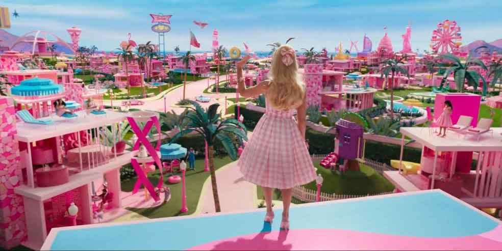 Imagem do filme Barbie (2023). Margot Robbie está caracterizada como Barbie, de costas em primeiro plano, chegando à Barbieland, uma cidade toda cor de rosa. Ela ergue uma mão e acena.