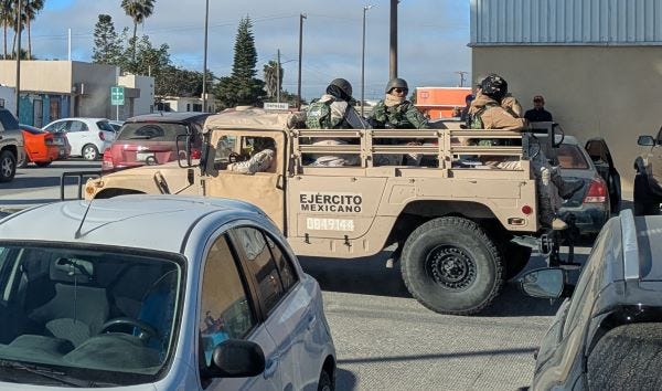 several soldiers in body armor with guns riding in the back of a tan colored military truck through the parking lot of a store.