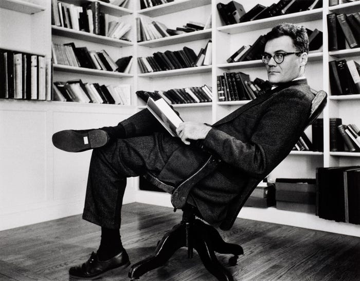 A tall, dark, suited man leans back in a chair reading a book in a study