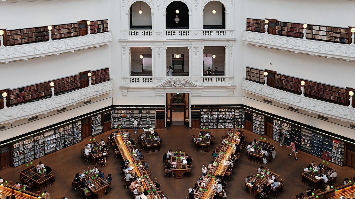 A photo of a grand library reading room with people working at desks and browsing books on shelves