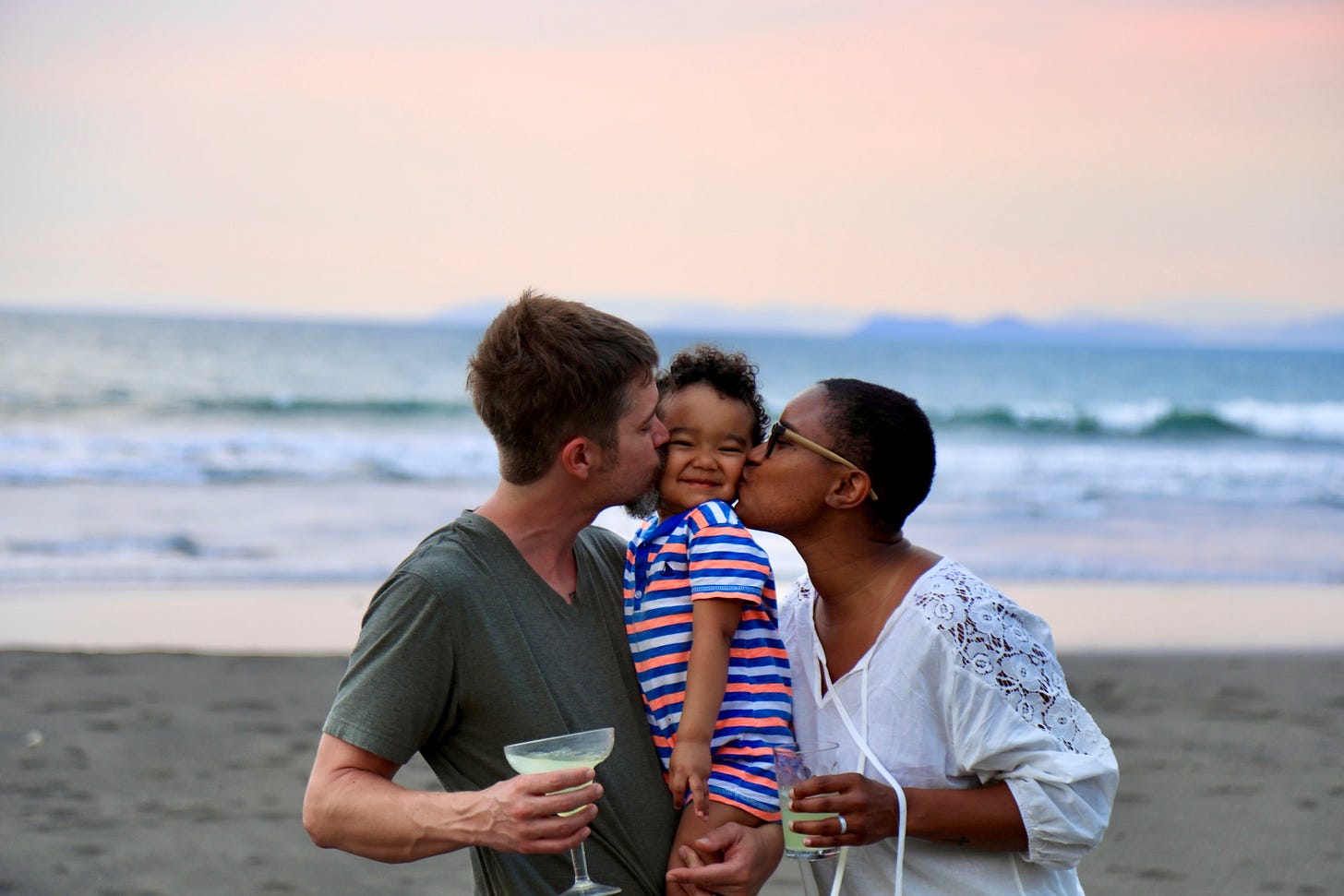 Mom and dad kissing toddler on the cheeks. The photo is on the beach in Costa Rica at sunset. 