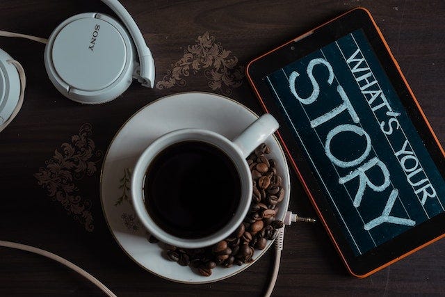 On a dark wood table sit a pair of headphones, a white cup filled with black coffee, the saucer full of coffee beans, and a tablet with a screen like a slate board reading “What’s Your Story”