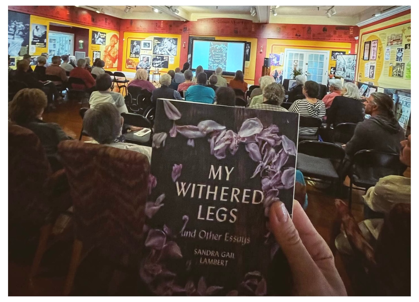 The view from the back of a large room with an audience seated in chairs. In the foreground is a hand holding up a copy of My Withered Legs and Other Essays by Sandra Gail Lambert