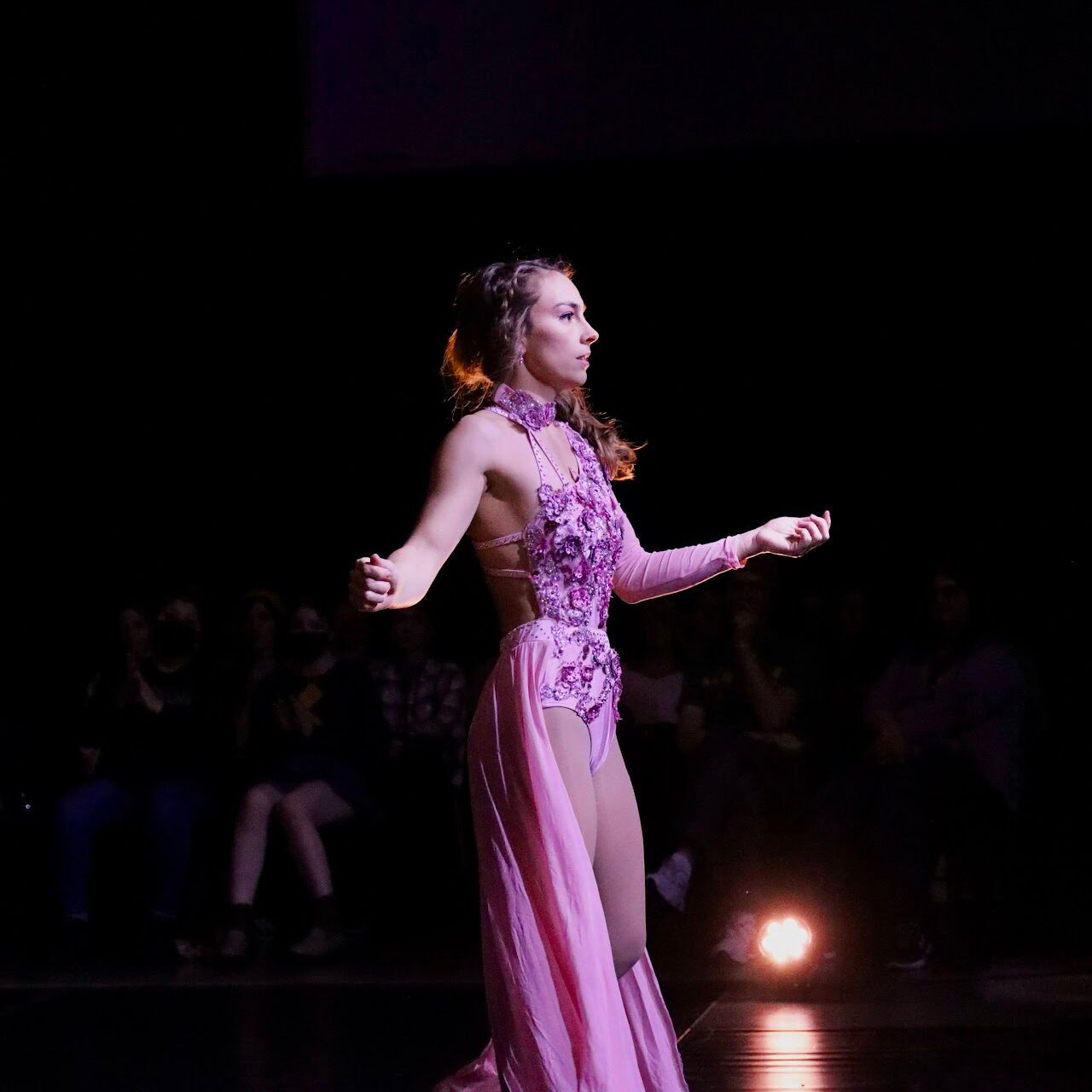 Alisha Rickman, wearing a flowing pink costume, performs on a dark stage illuminated by a spotlight. The audience, seated in the background, watches attentively as she gracefully moves. The costume features intricate detailing and a high neck.