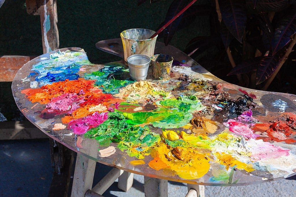 A large wooden paint pallete takes up most of the images and is covered in daubs of paint creating a rainbow of colours, you can see the energy of the way the artist has mixed the paint in the brush strokes on the surface. 