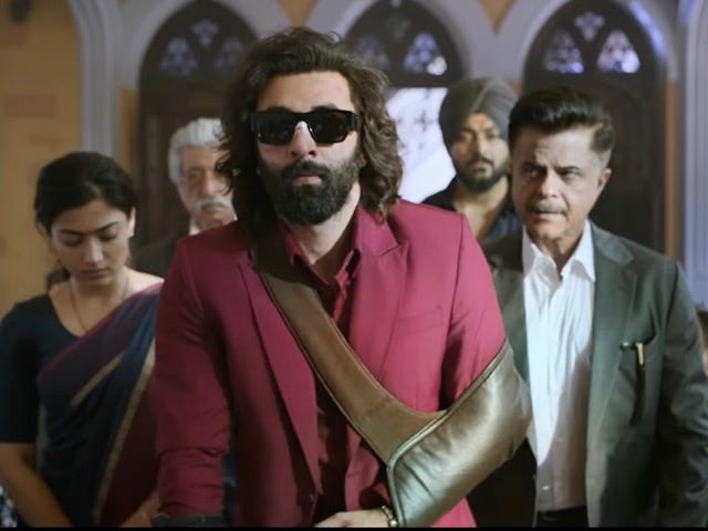 A still from Hindi film, ‘Animal’. A bearded and long-haired (Ranbir Kapoor) sporting sunglasses and a maroon suit and his left hand in a sling. (Anil Kapoor) and a few other people in the background.