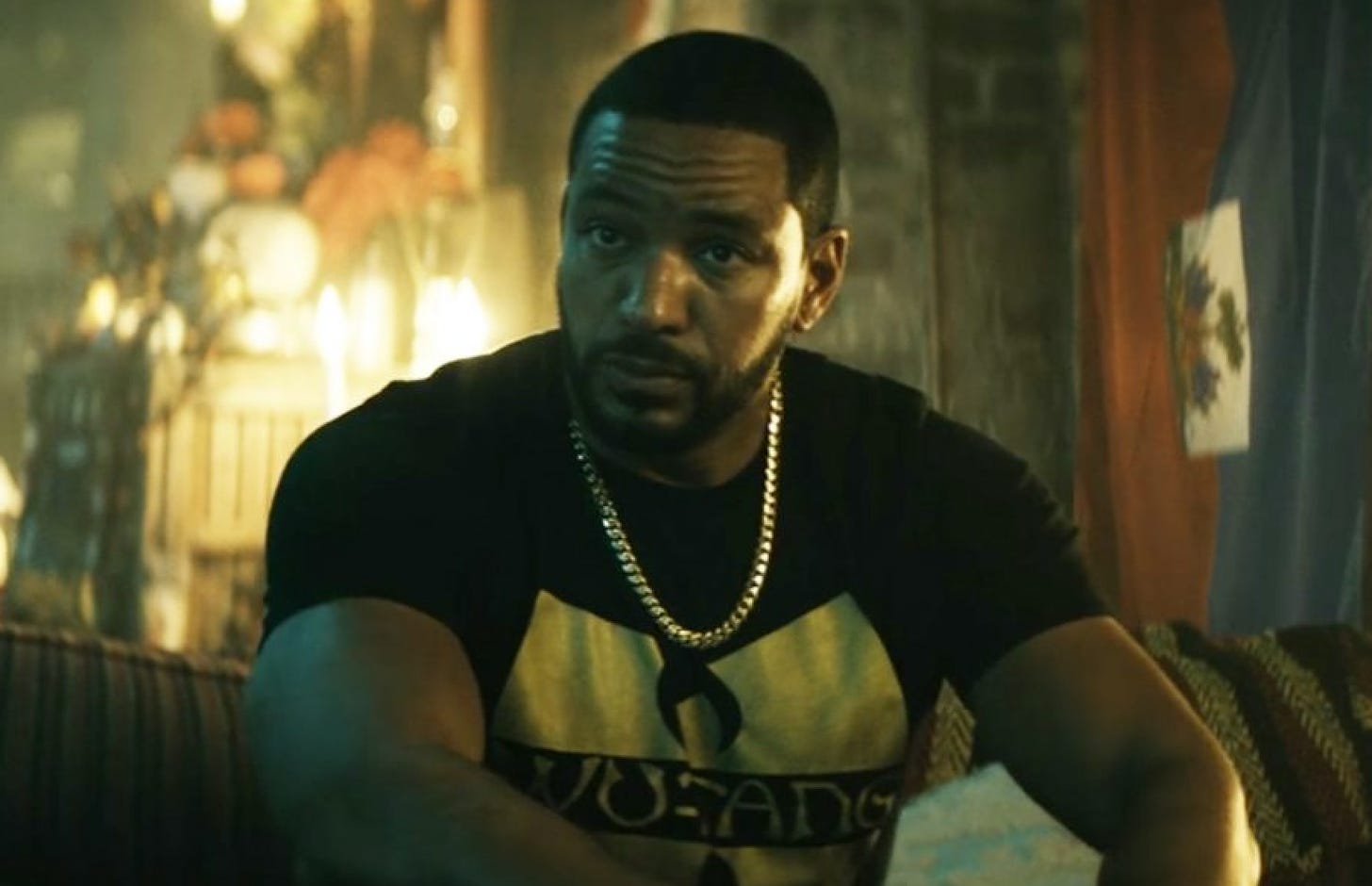 Laz Alonso as Mother's Milk" in "The Boys". He is sat on a couch wearing a Black tshirt with a concerned look on his face