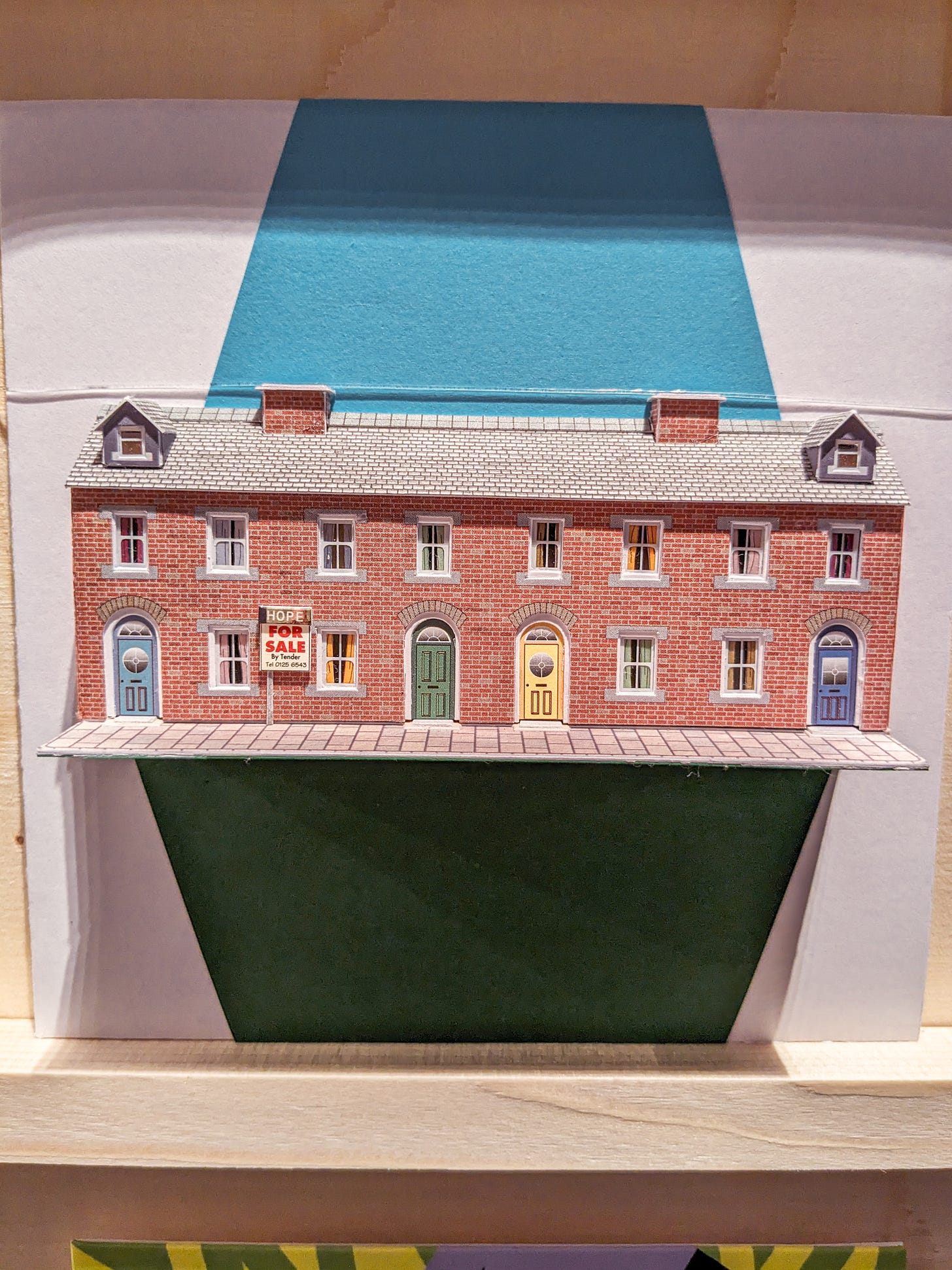 A 3D row of terraced houses made from card. There is a sign next to one of the windows that says "Hope for sale"