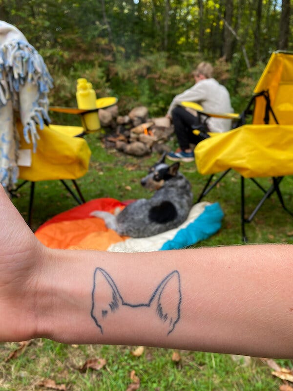 In the foreground, I hold up my right wrist showing a tattoo of Scout's ears — in the background, Scout and Sean sit around a fire