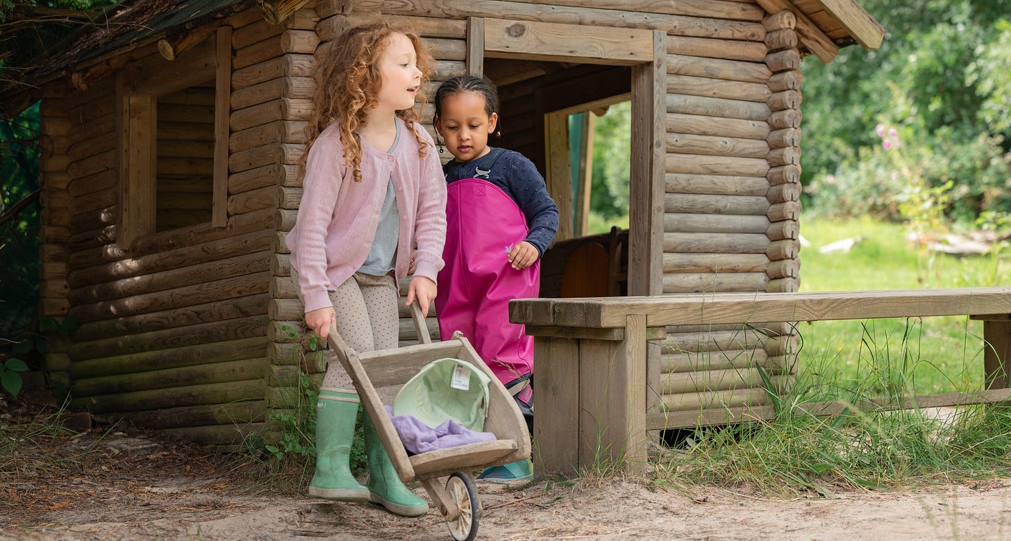 Two children playing outside with a wheelbarrow. There is a playhouse behind them, in a woodland setting