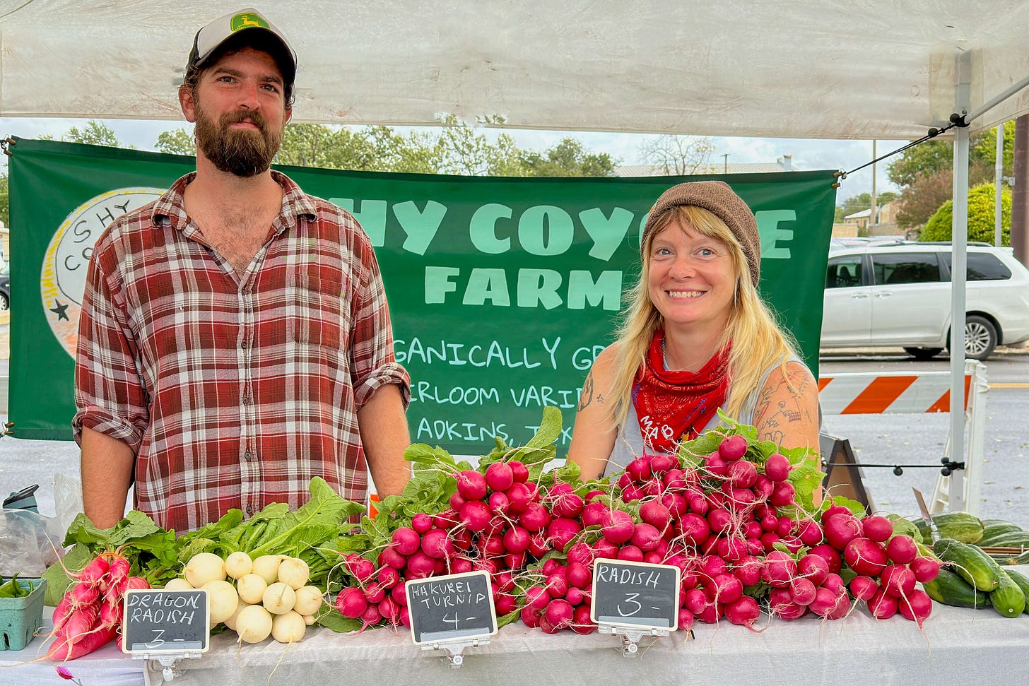 Zwei junge Farmer stehen hinter einem Marktstand auf einem Bauernmarkt in den USA und verkaufen frisches, biologisch angebautes Gemüse. Der Mann auf der linken Seite trägt ein kariertes Hemd und eine Baseballkappe, die Frau auf der rechten Seite hat blonde Haare, trägt eine Mütze und ein rotes Halstuch. Vor ihnen liegen Bündel von Radieschen, weissen Rüben und anderen Produkten, mit handgeschriebenen Preisschildern. Im Hintergrund ist ein grünes Banner mit dem Namen ihrer Farm sowie geparkte Autos zu sehen. Zwei junge Farmer stehen hinter einem Marktstand auf einem Bauernmarkt in den USA und verkaufen frisches, biologisch angebautes Gemüse. Der Mann auf der linken Seite trägt ein kariertes Hemd und eine Baseballkappe, die Frau auf der rechten Seite hat blonde Haare, trägt eine Mütze und ein rotes Halstuch. Vor ihnen liegen Bündel von Radieschen, weissen Rüben und anderen Produkten, mit handgeschriebenen Preisschildern. Im Hintergrund ist ein grünes Banner mit dem Namen ihrer Farm sowie geparkte Autos zu sehen.