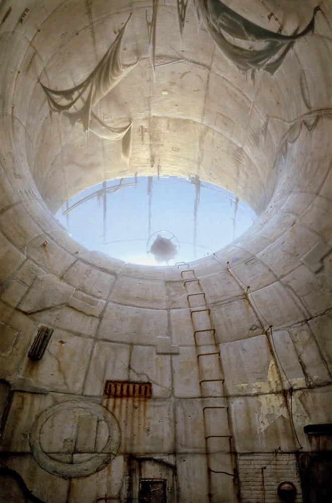 A low angled view follows the curve of concrete wall up a cavernous toroidal space. The structure at the upper edge of the curve drops off, opening the tunnel to blue sky. The contrast against pale concrete evokes the appearance of the human eye. At the lower center of the opening, a figure in a cloak holds a metal ring, which simultaneously looks like an iris while echoing the artist's sigil (the letters M and W combined inside a circle. Rebar set in concrete forms a ladder up the curving slope, further defined by its rust stained trail. Next to the ladder, a weathered five-pointed star is painted in yellow on the irregularly paneled concrete wall.