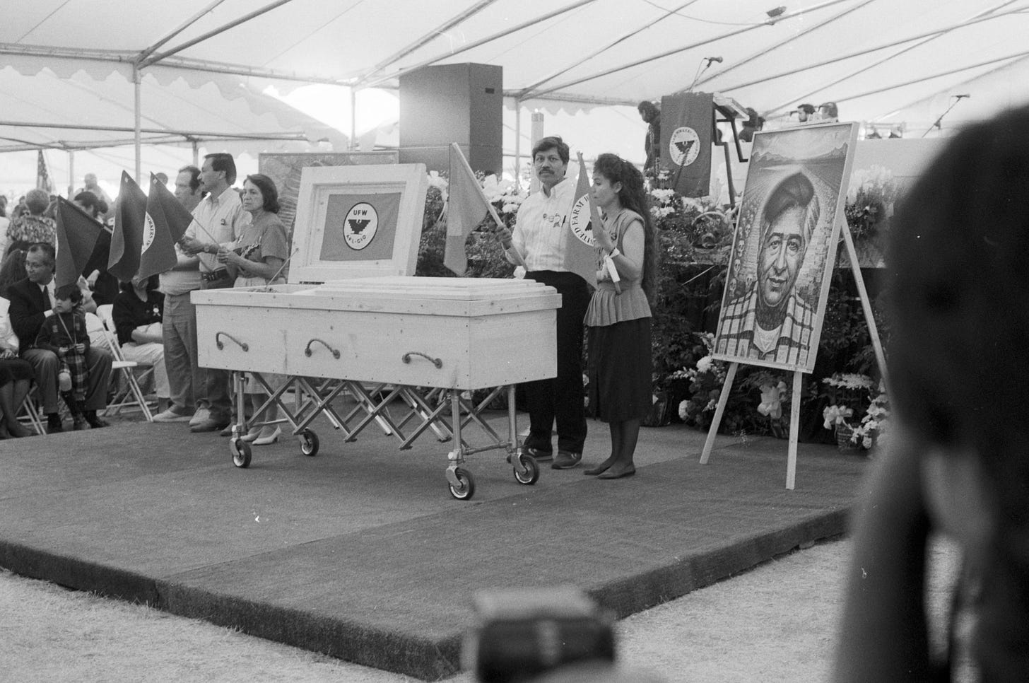 The body of César Chávez lies in repose during his funeral at Forty Acres. His body lies in a simple pine wood coffin made by his brother Richard Chávez at his request. A UFW banner adorns his coffin and the portrait of Chávez by Mexican artist Octavio Ocampo stands next to it. Dolores Huerta stands near the coffin holding a union flag. Photo by John Kouns © Tom and Ethel Bradley Center