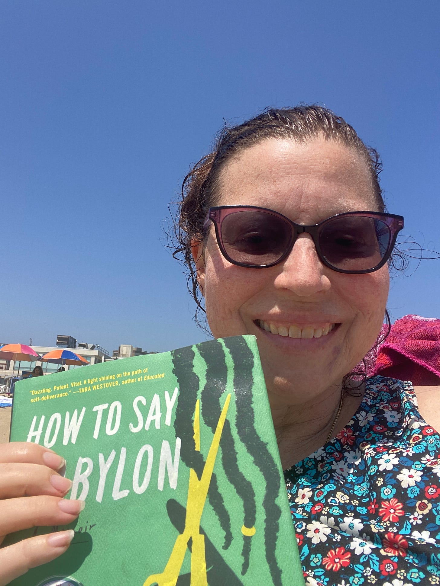 Gabrielle is a white skinned woman wearing sunglasses and sitting on the beach holding up a book. She smiles ar rge camera