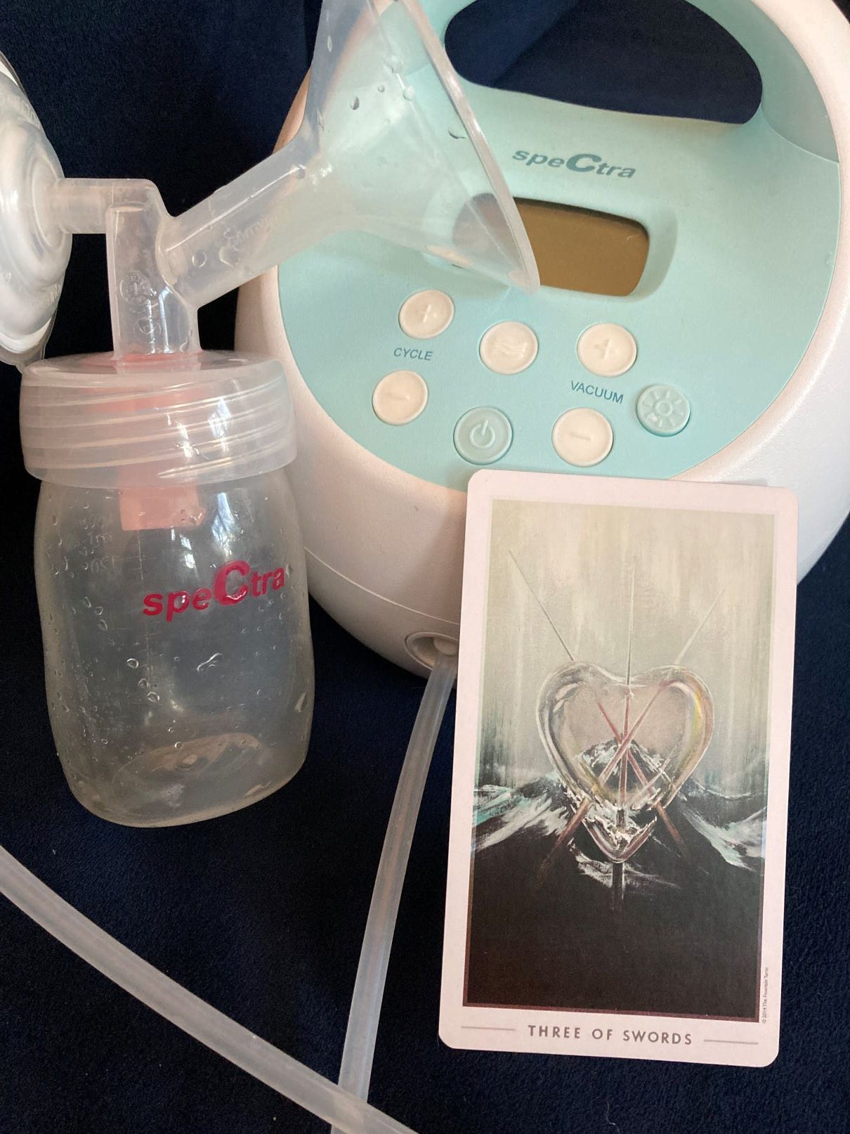 The tarot card Three of Swords sits next to a breast pump against a blue velvet background