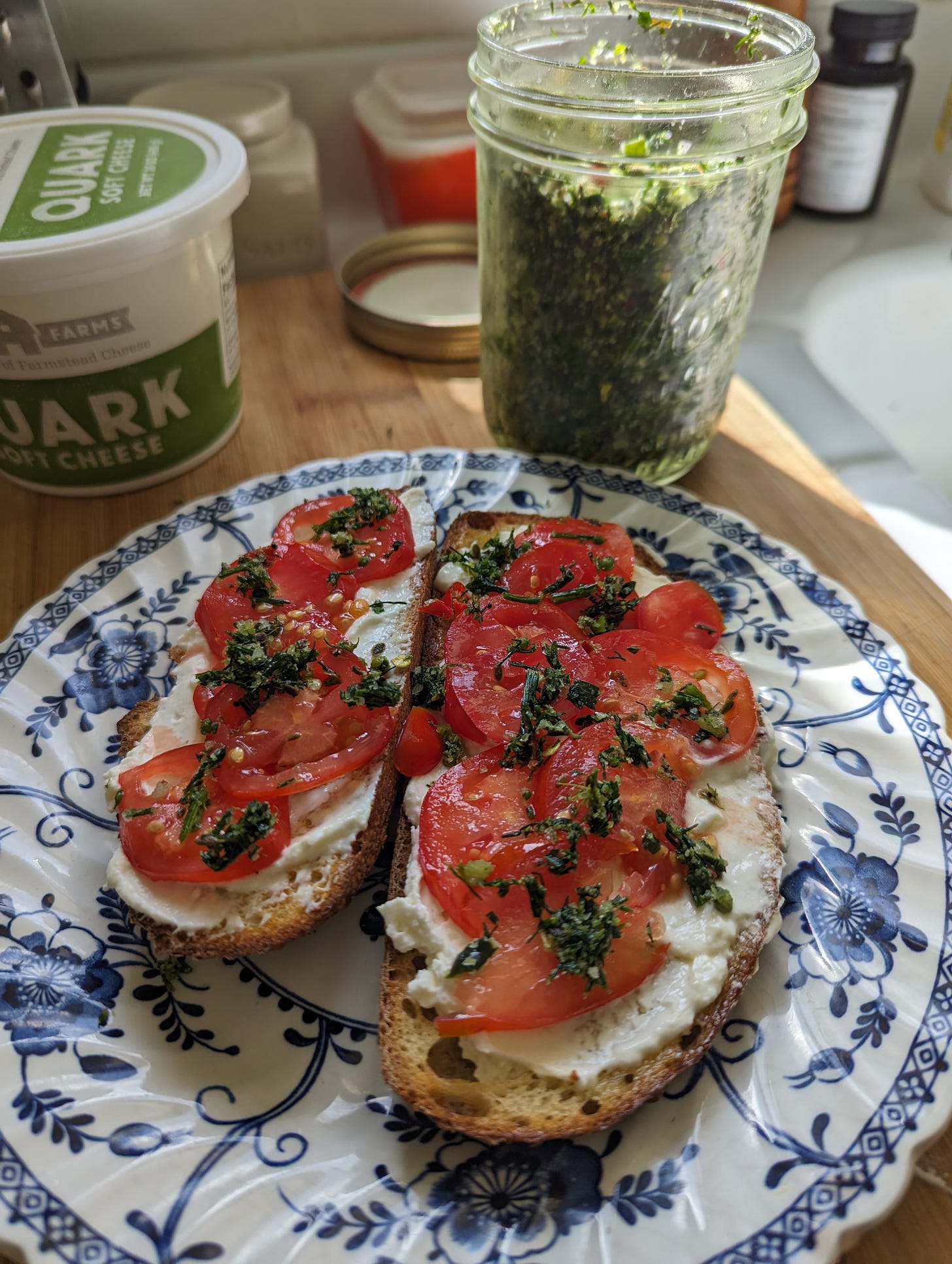 Jar of green salt and tub of Quark cheese in background. Foreground: blue and white plate with toast slices topped with quark, sliced tomatoes, and green salt. 