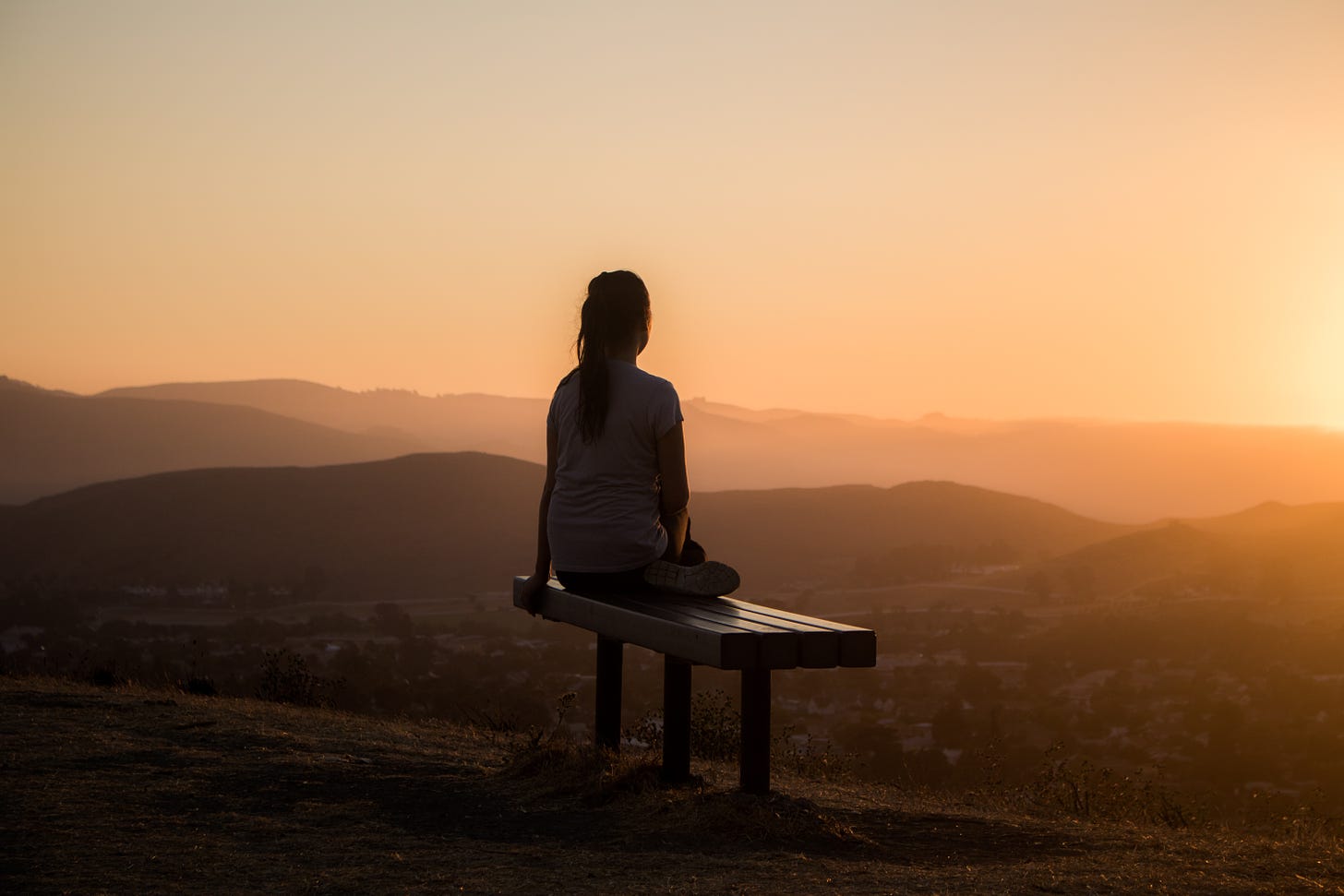 Silhouette of woman from behind, sitting on a bench watching a sunset from a hilltop