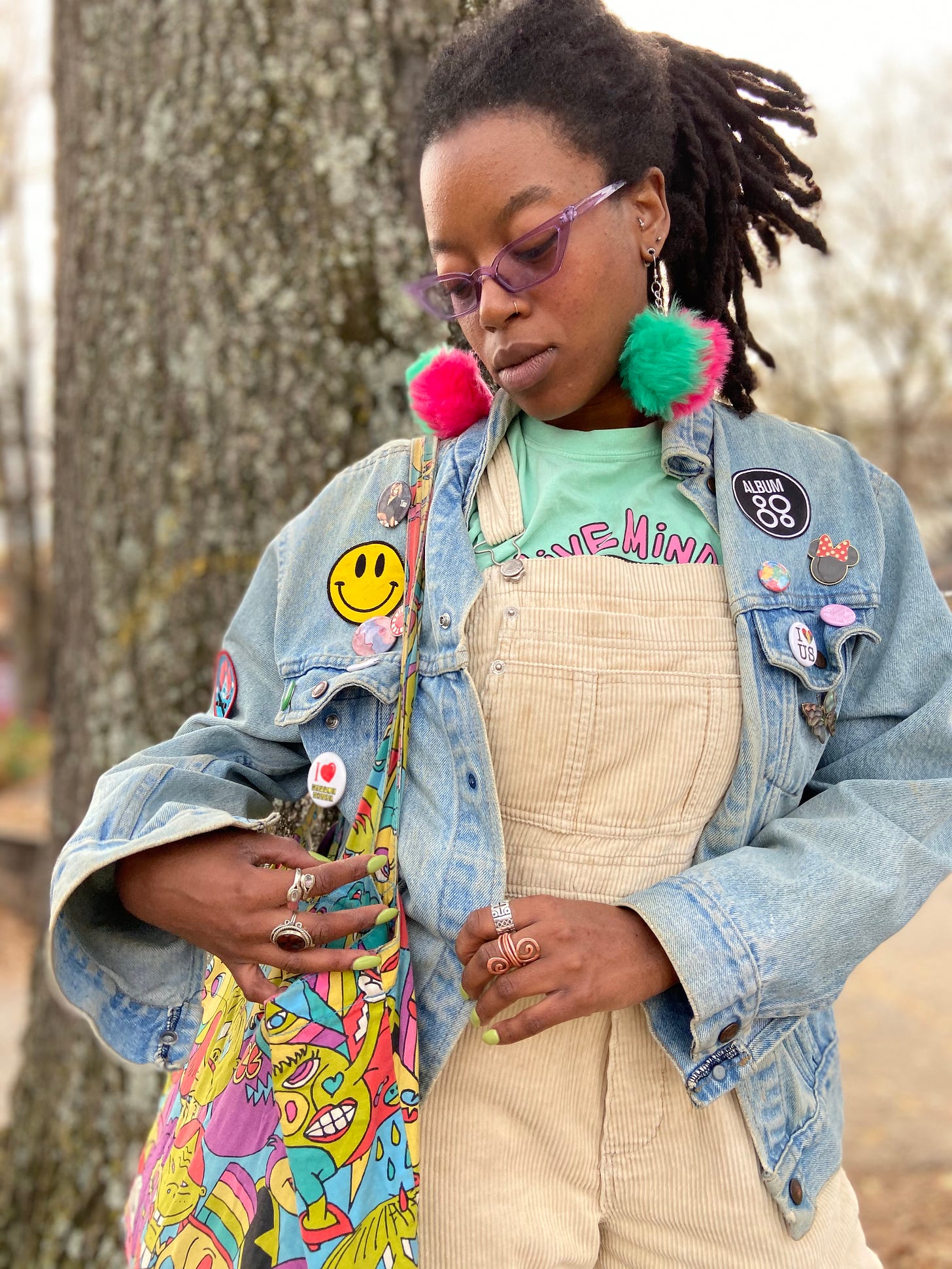 Tay, a Black person, poses with purple sunglasses, pom-pom earrings, tan overalls, a band tee, a denim jacket covered in pins, and a colorful tote bag.